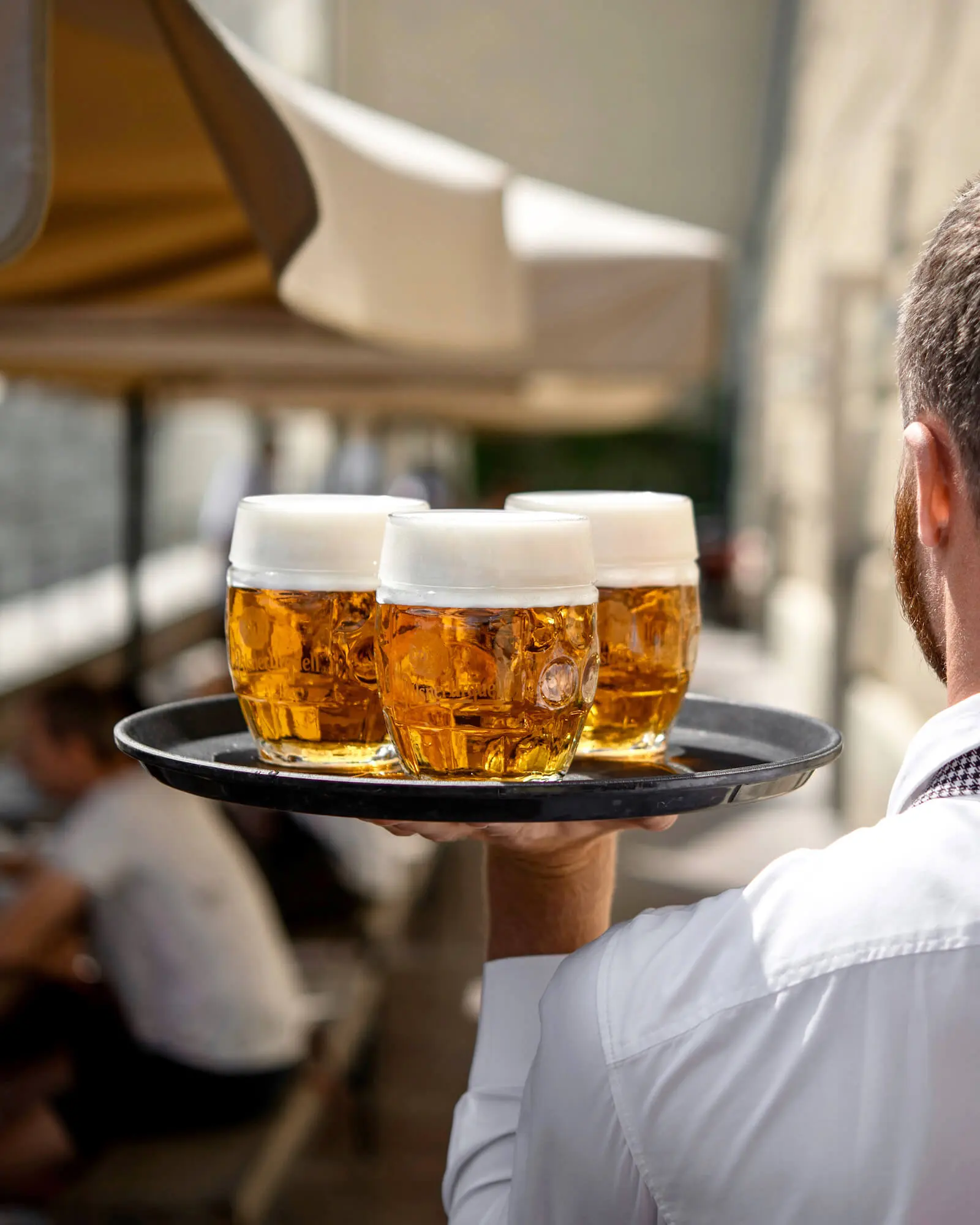 A waiter carrying a tray with three mugs of beer topped with thick foam on an outdoor terrace.